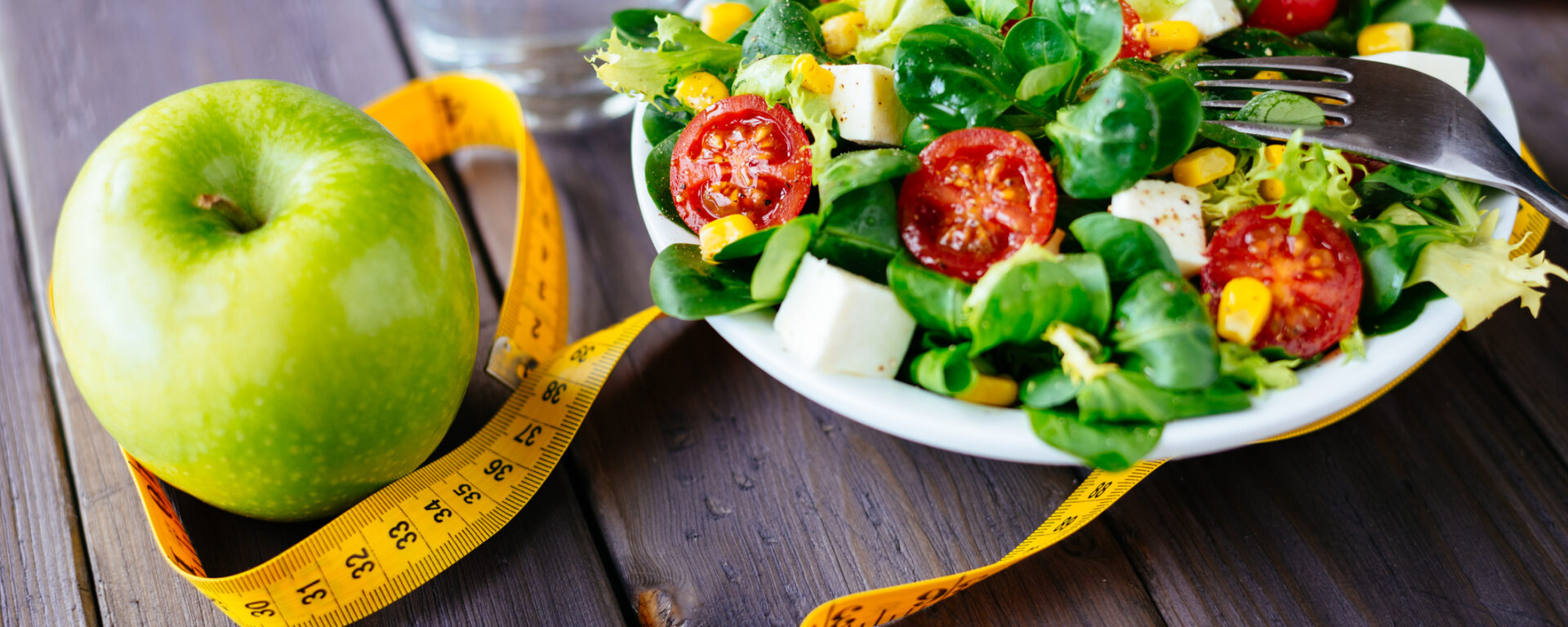 Fitness salad and apple fruit surrounded by measuring tape on rustic wooden table. Mixed greens, tomatos, diet cheese, olive oil and spices for healthy lifestyle concept.