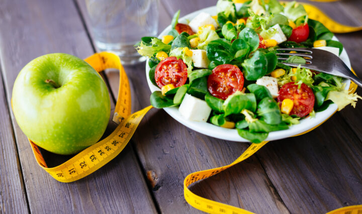 Fitness salad and apple fruit surrounded by measuring tape on rustic wooden table. Mixed greens, tomatos, diet cheese, olive oil and spices for healthy lifestyle concept.